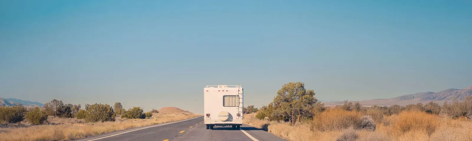 An RV driving down an open, two-lane highway through a vast desert landscape under a clear, bright blue sky.
