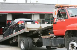 Red flatbed tow truck loading a dark sedan onto its tilted bed outside an auto repair shop