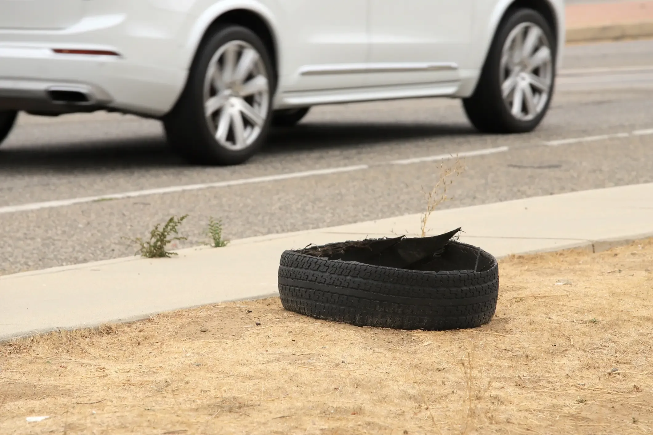 Shredded blown-out tire lying on a dry roadside shoulder as a white SUV passes by