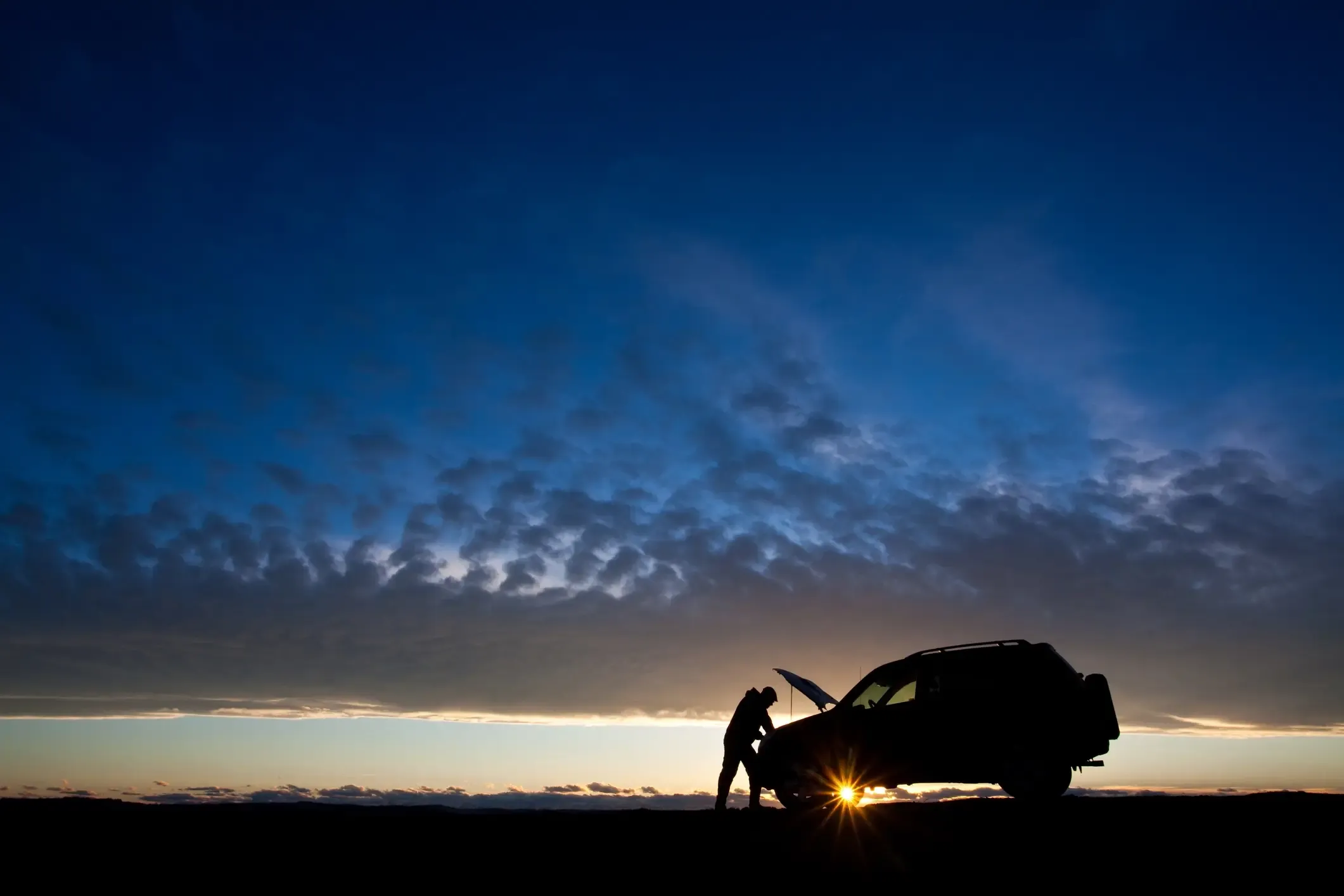 Silhouetted figure leaning over an SUV's open hood as the sun sets beneath a cloudy blue sky
