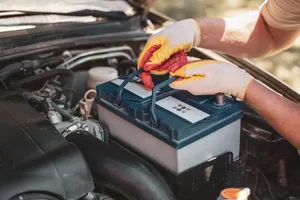 Gloved hands lifting a car battery into the engine bay by its carry strap during a battery replacement