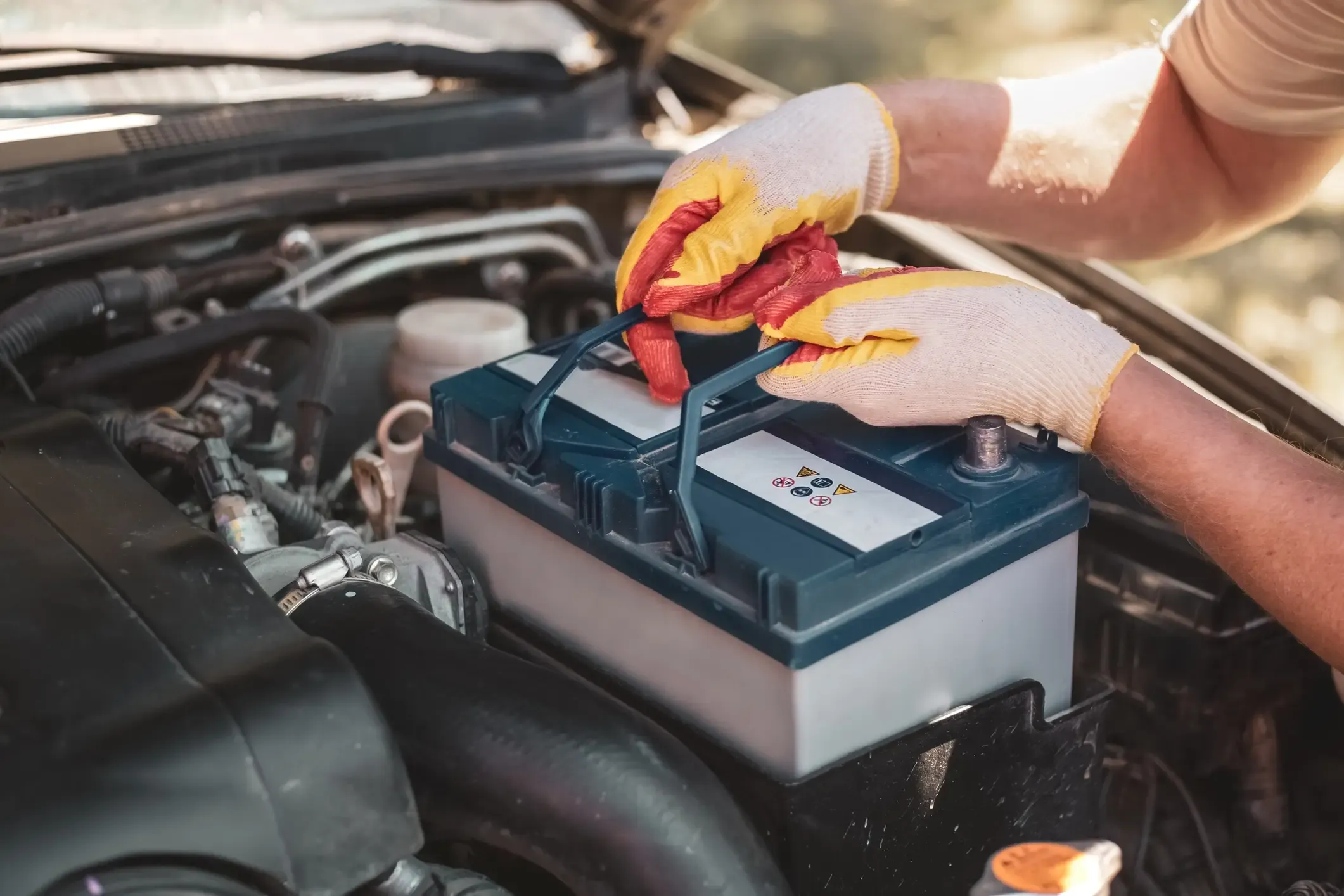 Gloved hands lifting a car battery into the engine bay by its carry strap during a battery replacement