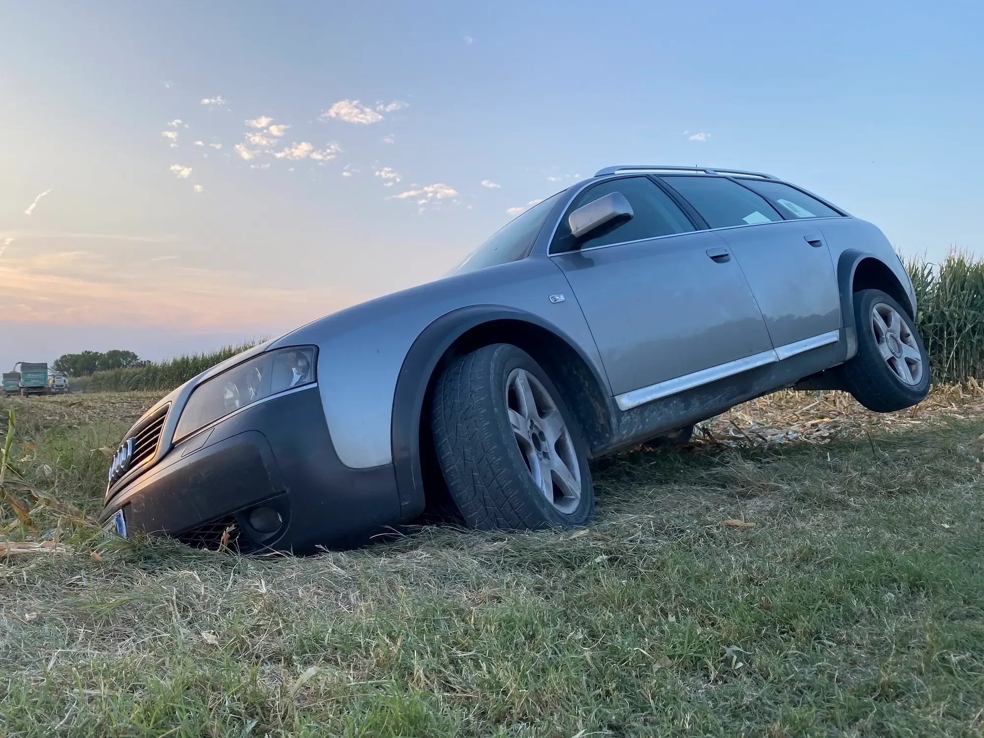 Silver car stuck nose-down in a roadside ditch by a cornfield, in need of recovery at dusk