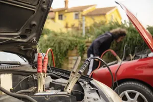 Red and black jumper cable clamps attached to a car battery as a person jump-starts a red car nearby.