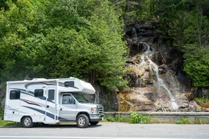 White Class C motorhome parked beside a roadside waterfall and lush green forest