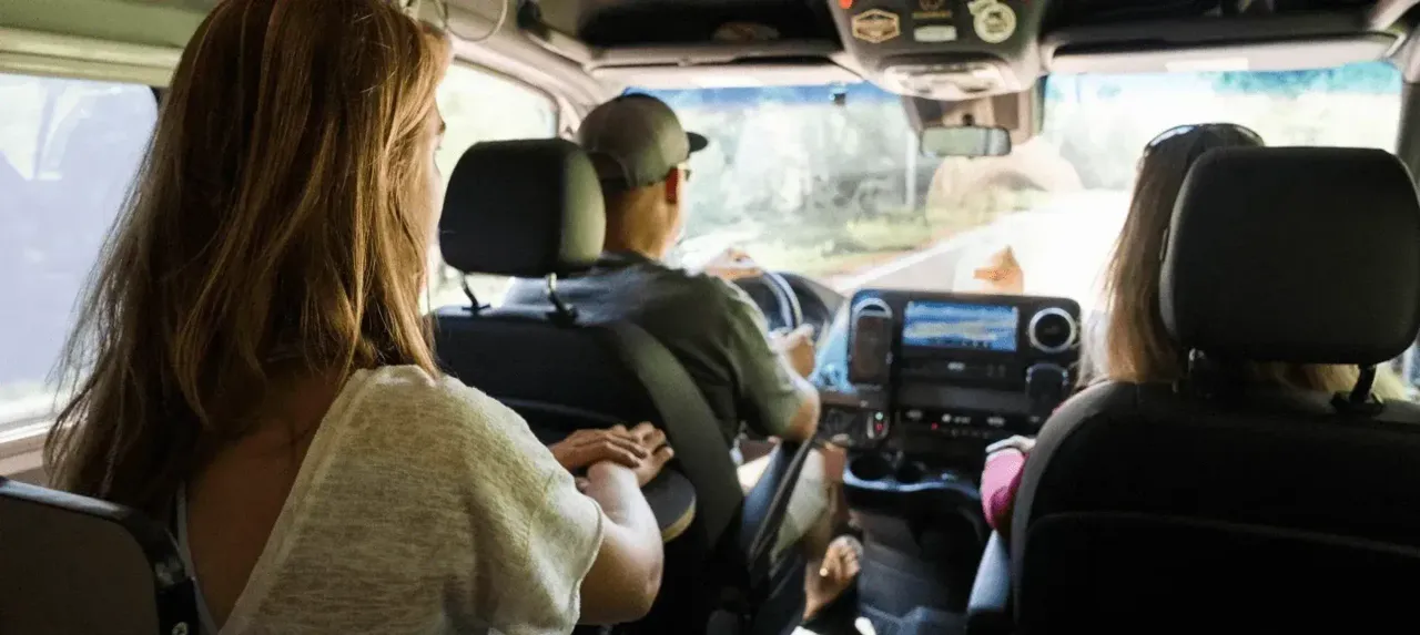 Three people ride in a van driving down a road on a sunny day.