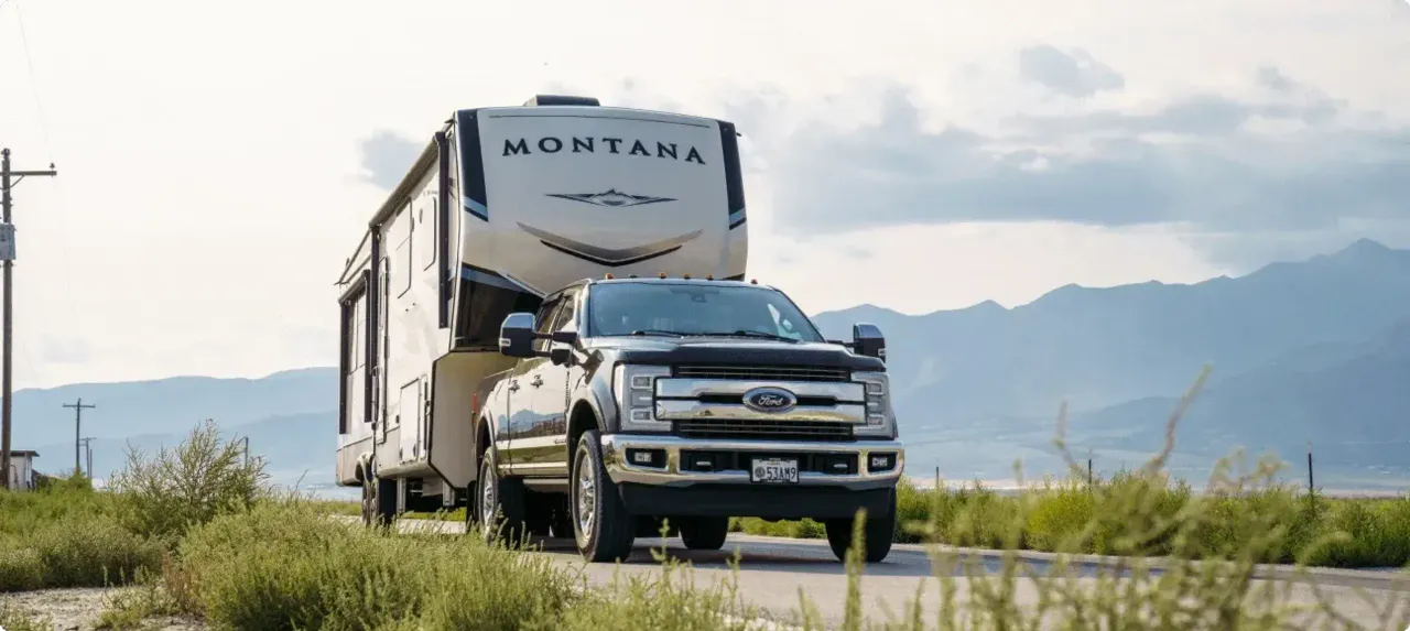 A truck tows a Montana fifth week along a road in with a backdrop of tall rocky mountains.