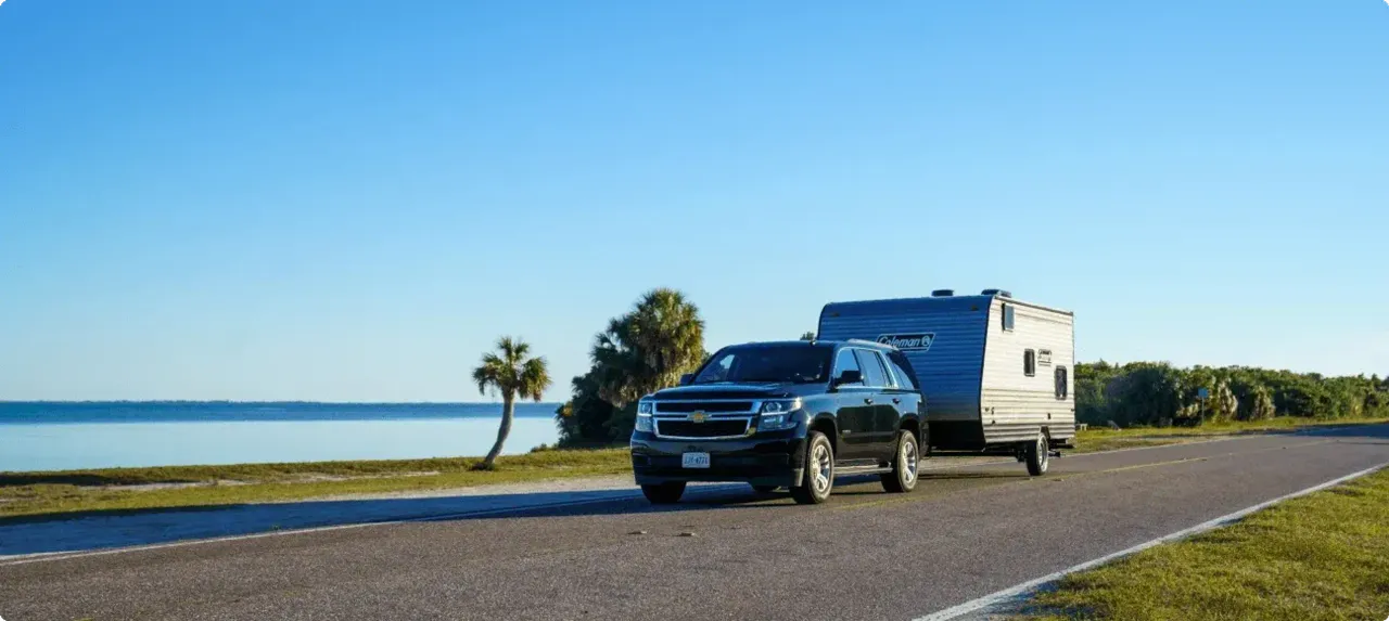An SUV pulls a trailer alongside a coastal waterway with a palm tree in the background.