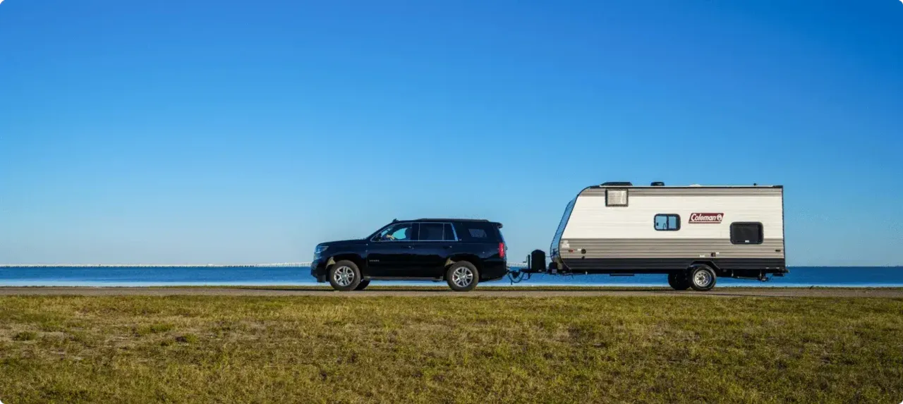 An SUV pulls a Coleman trailer along a road with a coastal waterway in the background.
