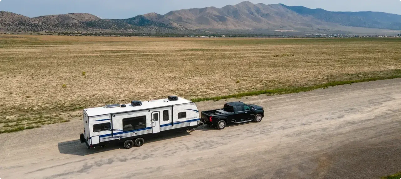 A truck tows a fifth wheel in front of a wide dusty field with tall rocky mountains in the background