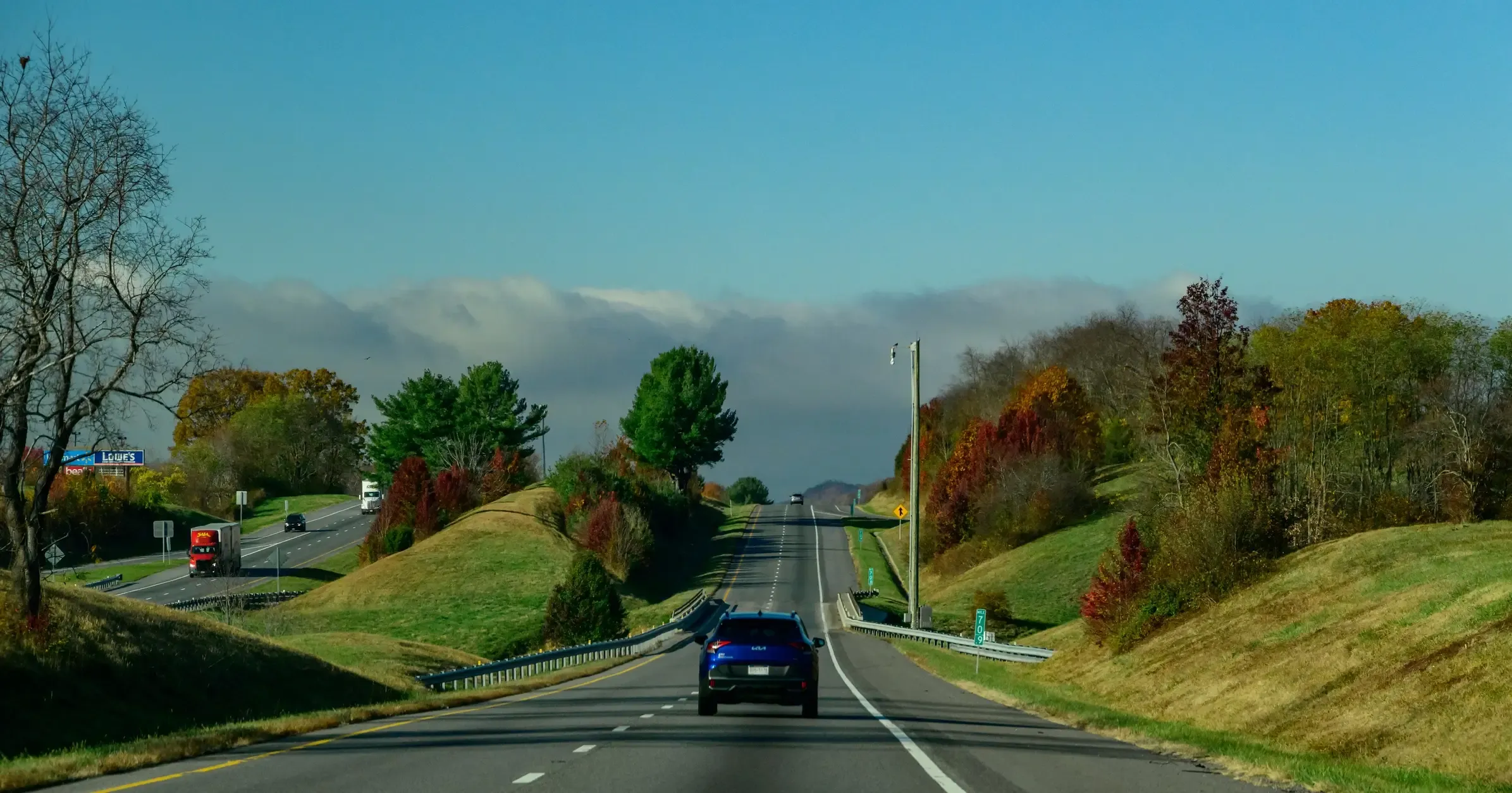 Blue SUV driving a divided highway through rolling hills with autumn foliage and distant fog