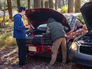 New driver learning to use jumper cables while an adult connects a dead battery under the hood.