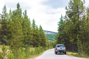 Pickup truck cruising a scenic two-lane road lined with evergreens in the mountains.