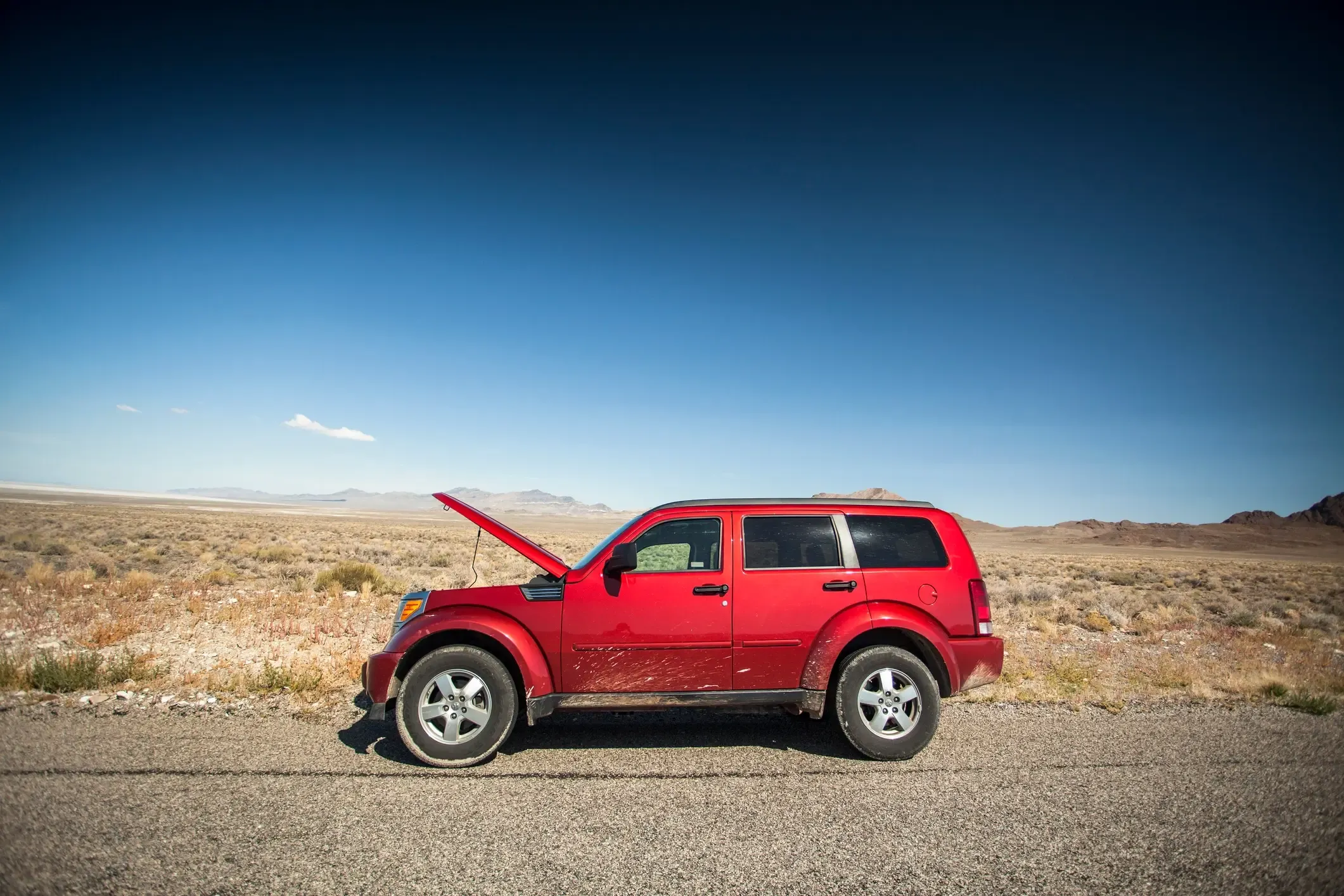 Red SUV with its hood up, broken down on a remote desert roadside with mountains in the distance