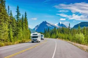 An RV on the road with trees on the side of the road and mountains off in the distance. 