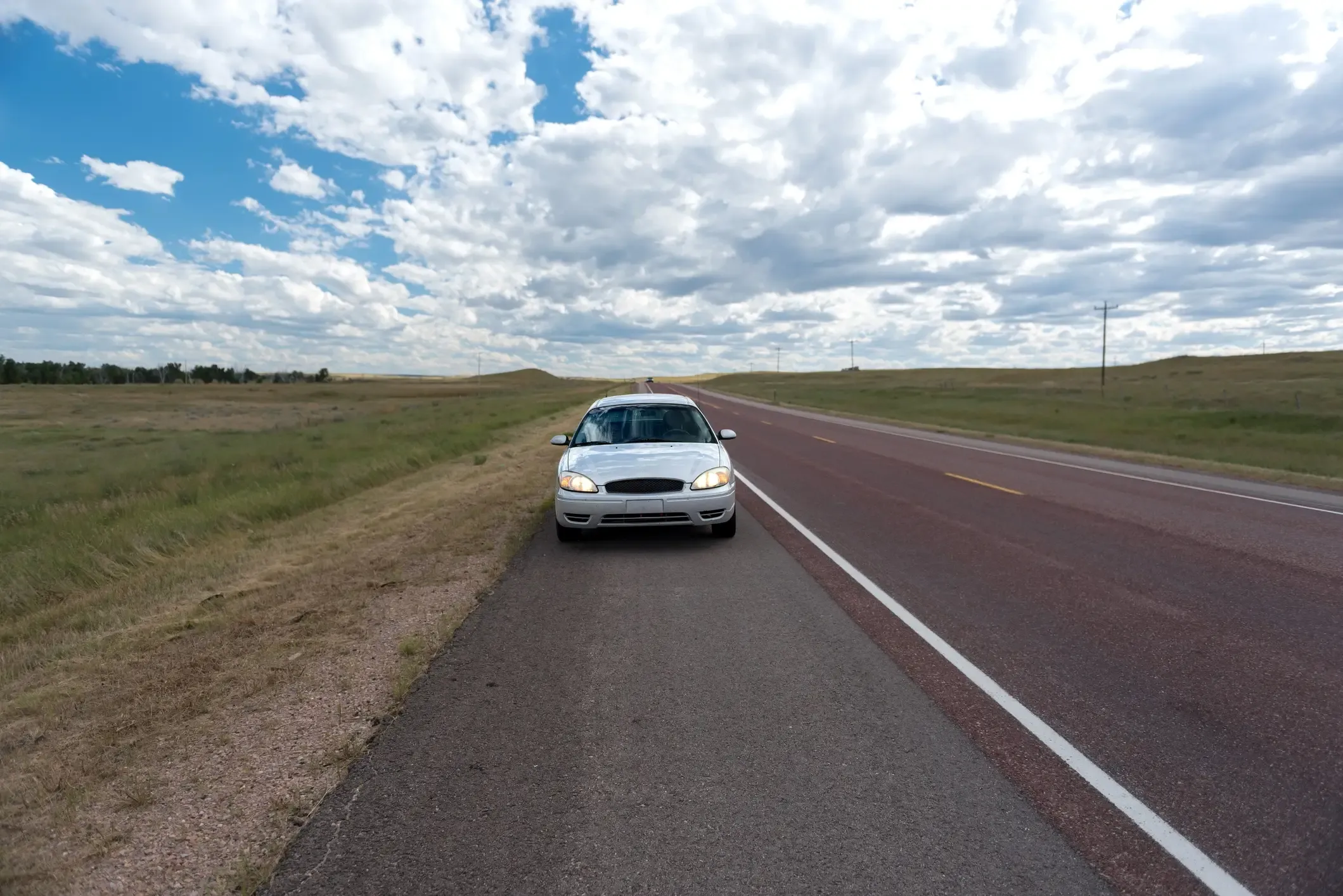 White sedan pulled over on the shoulder of an empty rural highway under a cloudy sky