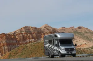 Silver Class C motorhome parked on a desert road in front of dramatic red rock cliffs