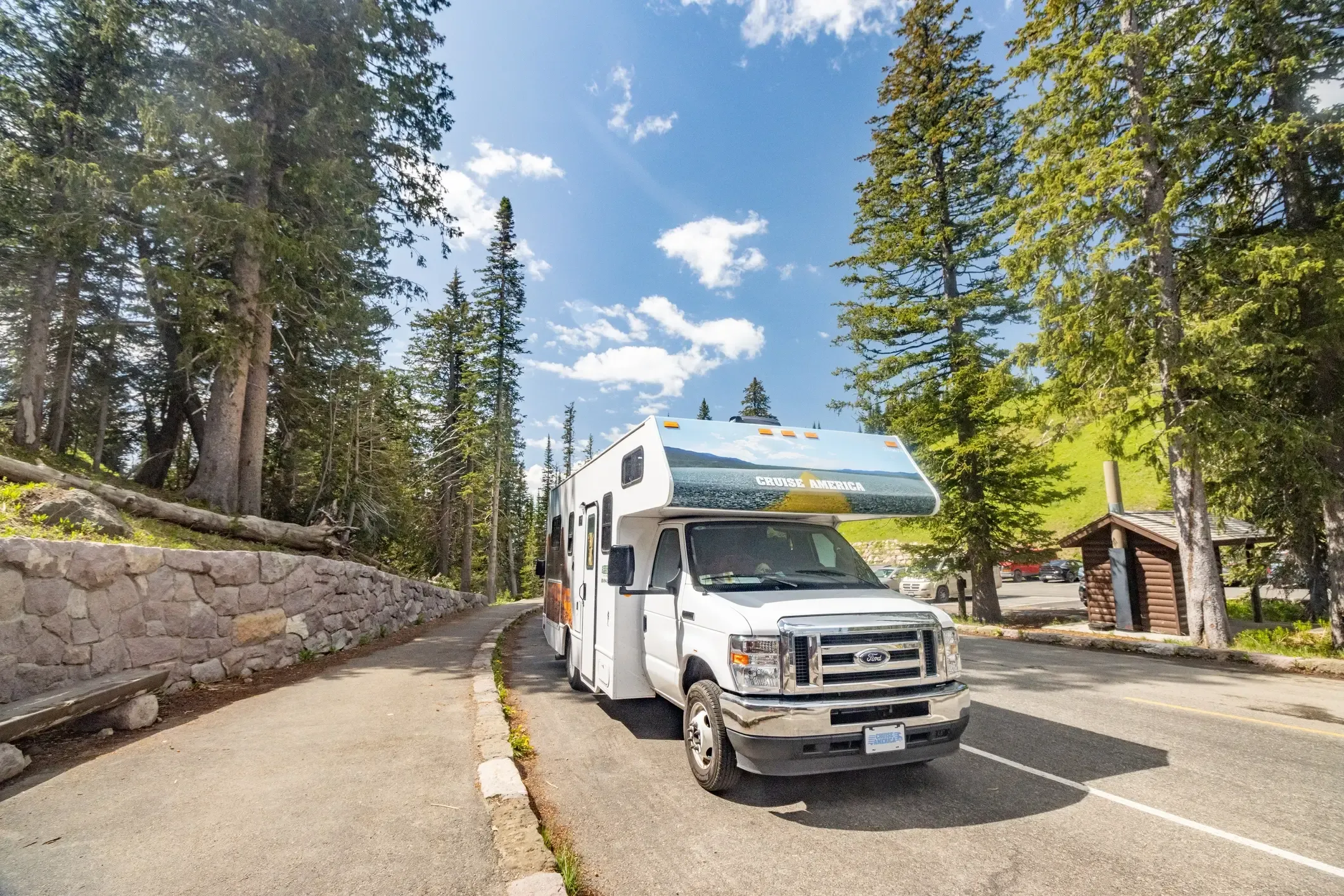 Class C rental motorhome driving a forest road through a mountain park lined with tall pines